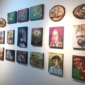 Op de afbeelding: Een galeriewand met een verzameling rechthoekige canvasafdrukken en cirkelvormige kunstwerken op houten schijven. De kunstwerken bevatten diverse popcultuurillustraties, portretten en grafische ontwerpen, gerangschikt in een raster op een witte muur.