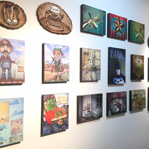 Op de afbeelding: Een verzameling van diverse rechthoekige en ronde wandkunstwerken met uiteenlopende illustraties, waaronder personages zoals Mario, Link en Batman, naast in hout gebrande ontwerpen, allemaal samen tentoongesteld op een witte muur.