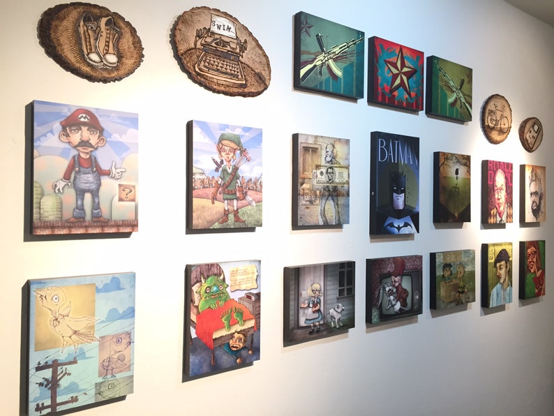Op de afbeelding: Een verzameling van diverse rechthoekige en ronde wandkunstwerken met uiteenlopende illustraties, waaronder personages zoals Mario, Link en Batman, naast in hout gebrande ontwerpen, allemaal samen tentoongesteld op een witte muur.