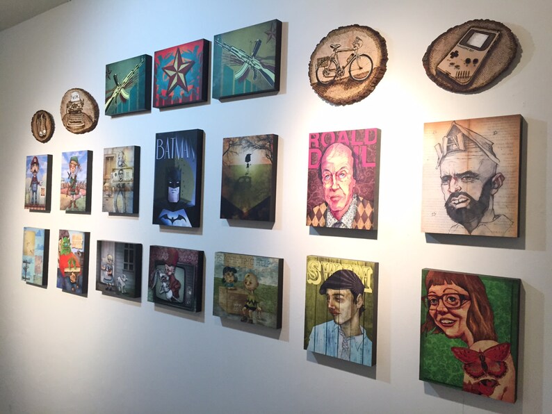 K&ouml;nnte beinhalten: Eine Galeriewand mit einer Sammlung von rechteckigen Leinwanddrucken und kreisf&ouml;rmigen Holzscheiben-Kunstwerken. Die Kunstwerke umfassen verschiedene Popkultur-Illustrationen, Portr&auml;ts und Grafikdesigns, die in einem Raster an einer wei&szlig;en Wand angeordnet sind.
