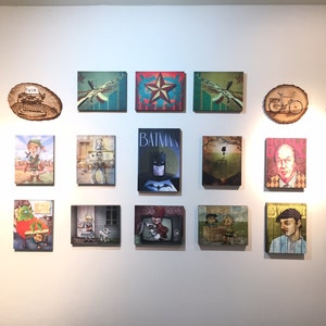 Op de afbeelding: Een galerijwand met eenentwintig kunstwerken, waaronder rechthoekige canvasprints en ronde houten schijven. De collectie toont diverse popcultuurfiguren, gestileerde portretten en grafische illustraties in een verscheidenheid aan kleuren.