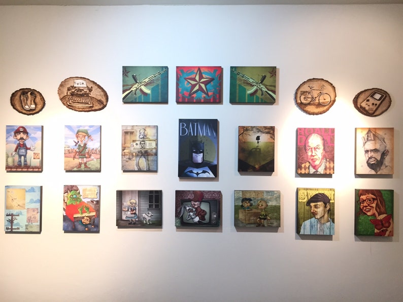 Op de afbeelding: Een galerijwand met eenentwintig kunstwerken, waaronder rechthoekige canvasprints en ronde houten schijven. De collectie toont diverse popcultuurfiguren, gestileerde portretten en grafische illustraties in een verscheidenheid aan kleuren.