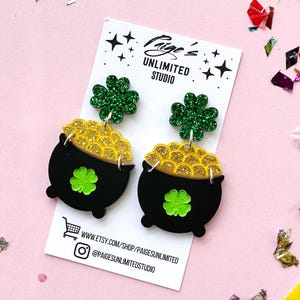 May include: Black and gold pot of gold earrings with green shamrock accents. The earrings have a glittery green shamrock at the top and a black pot filled with gold coins. The earrings are on a white card with the text "Paige's Unlimited Studio".