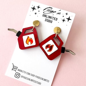 May include: Red and white earrings shaped like gas cans, featuring a flame design. The earrings have gold-colored studs and silver-colored nozzles. The earrings are displayed on a white card with the text "Paige's Unlimited Studio".