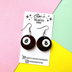 May include: A pair of dangle earrings featuring black circular charms with a white center and the number 8, resembling billiard balls. The earrings are attached to a white display card printed with the text Paige's Unlimited Studio.