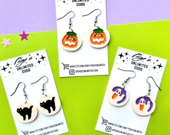 Könnte beinhalten: Drei Paar Halloween-Ohrringe auf weißen Karten. Ein Paar zeigt orangefarbene Kürbisse, ein weiteres schwarze Katzen und das letzte lila Geister. Jede Karte trägt den Text "Paige's Unlimited Studio" und Social-Media-Informationen.