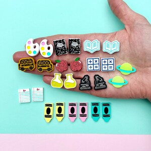May include: A selection of colourful school-themed charms, including paint palettes, school buses, apples, notebooks, calculators, planets, beakers, microscopes, A+ papers, and crayons. The charms are in various colours and designs, ideal for crafting or jewellery making.