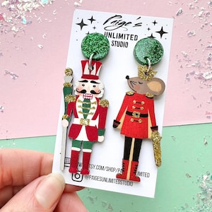 May include: A pair of festive earrings featuring a nutcracker and a mouse soldier design. The earrings have green glitter tops and are held against a pink and green background. The nutcracker is in a red, white, and green uniform, while the mouse is in a red uniform.