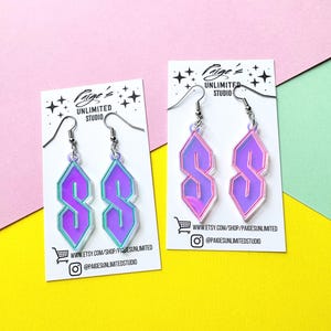 May include: Two pairs of dangle earrings featuring the iconic geometric S shape in iridescent purple and teal acrylic. Each pair is mounted on a white display card labelled Paige's Unlimited Studio with social media handles and a URL.