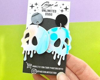 May include: A pair of skull-shaped earrings with a white, blue, and tan marbled design. The earrings have a dripping effect at the bottom and are attached to a black and silver stud. The earrings are displayed on a black card with the text "Paige's Unlimited Studio".