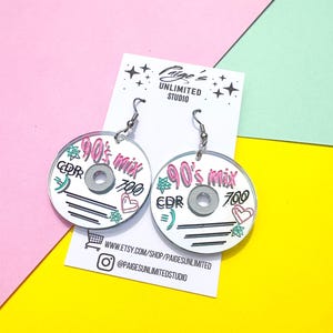 May include: A pair of circular acrylic earrings shaped like compact discs, featuring the text 90's mix, CDR, and 700 in black, pink, and teal ink. The earrings are attached to a white display card labeled Paige's Unlimited Studio with social media info.