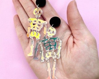 May include: A pair of iridescent skeleton earrings. Each earring features a skeleton design with a black circular stud at the top. The skeletons have a colorful, translucent appearance with yellow, green, and orange hues. The earrings are held in a hand against a pink background.