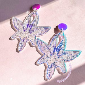 May include: A pair of iridescent earrings shaped like angels with wings. The earrings are made of clear acrylic and have a holographic finish. The wings are detailed with many eyes. The earrings are hanging from purple posts.