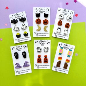 May include: A collection of Halloween-themed stud earrings on white cards. Designs include bats, ghosts, pumpkins, skulls, coffins, witch hats, fangs, candy corn, and chocolate bars. The cards have the text "Paige's Unlimited Studio" and social media handles.