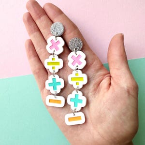 May include: A pair of long dangle earrings featuring silver glitter circular studs and four stacked white geometric shapes with cutouts. The cutouts contain bright pink, yellow, turquoise, and orange mathematical symbols including plus, minus, and division signs.