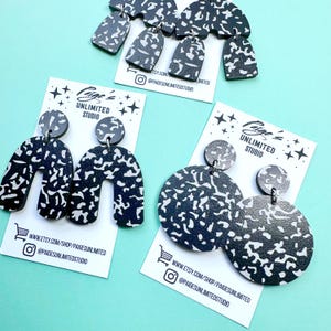 May include: A collection of handmade earrings in various shapes, including arches, circles, and teardrops. The earrings are black with a white speckled pattern. Each pair is displayed on a white card with the text "Paige's Unlimited Studio".