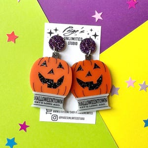 May include: A pair of dangle earrings featuring orange acrylic jack-o'-lantern pumpkins with black glitter details. Each pumpkin sits atop a white rectangular base printed with the text HALLOWEENTOWN EST'D LONG AGO. The posts are purple glitter circles.