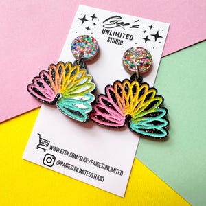 May include: A pair of dangle earrings featuring circular studs with multicolored glitter and fan-shaped pendants with a black base and a rainbow-colored gradient design. The earrings are displayed on a white card labeled Paige's UNLIMITED STUDIO.