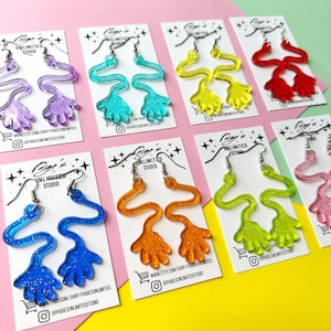 May include: A collection of dangling earrings shaped like cartoon hands with long, wavy arms, each pair featuring a distinct glittery colour including purple, teal, yellow, red, blue, orange, and lime green, all displayed on individual white branded cards.