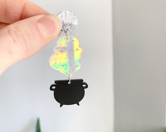 May include: A pair of dangle earrings featuring a silver glitter disc, a holographic cloud shape with stars, and a black cauldron. The earrings are designed with a witchy or Halloween theme.