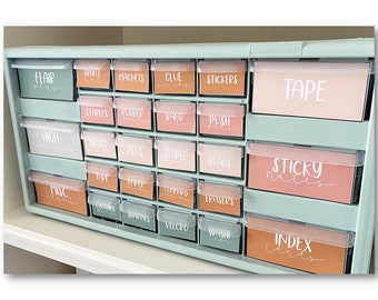 May include: A light blue drawer unit with labels for various school supplies. The labels are in a peach, pink, and brown color scheme. The labels include "Flair Pens", "White Out", "Magnets", "Glue Sticks", "Stickers", "Tape", "Staples", "Rubber Bands", "Band Aids", "Push Pins", "Highlighters", "Binder", "Pens", "Staple", "Brads", "Sticky Notes", "Misc", "Tape", "Paper Clips", "Command Strips", "Erasers", "Clothes Pins", "Sharpies", "Velcro", "Wash Tape", and "Index Cards".