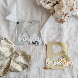 May include: Two mirrored acrylic name tags. One silver tag with the letter 'K' and the name 'Kinsley', and a gold tag with the letter 'B' and the name 'Brooklyn'. Both have white ribbon. Decorative flowers are in the background.
