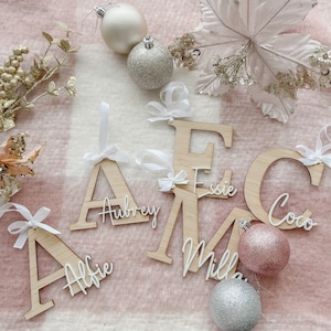 May include: Wooden ornaments shaped like the letters A, E, M, and C, with the names Alfie, Aubrey, Essie, Milla, and Coco written on them. The ornaments are decorated with white ribbon bows and are hanging on a pink and white blanket.