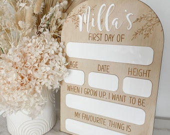 Puede incluir: Un letrero de madera con un arco blanco y el texto "Milla's First Day Of" en la parte superior. El letrero tiene espacios en blanco para la edad del niño, la fecha, la altura, lo que quiere ser cuando sea mayor y su cosa favorita.