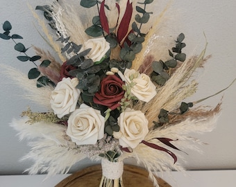 May include: A bridal bouquet featuring cream-colored and burgundy roses, accented with eucalyptus, pampas grass, and other dried foliage. The stems are wrapped with a cream-colored lace ribbon. The bouquet is arranged on a wooden surface.