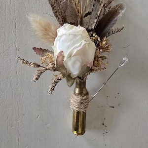 May include: A white rose with dried grasses and feathers arranged in a brass bullet casing. The casing is wrapped with twine.