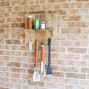 May include: A wooden shelf with hooks attached to a brick wall. The shelf holds three glass jars with lids and a spice jar. There are four grilling tools hanging from the hooks.