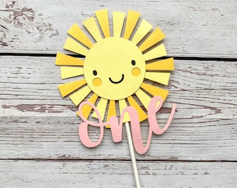 May include: A yellow paper sun with a smiling face and pink rays, with the word "one" in pink script on a white stick.