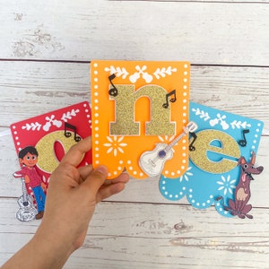 May include: A colorful banner with the letters "one" in gold glitter. The banner is decorated with musical notes, a guitar, a character from the movie Coco, and a dog. The banner is perfect for a first birthday party.