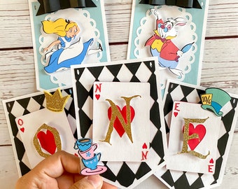 May include: A colorful banner with a checkered pattern and cut-out images of Alice in Wonderland characters. The banner reads "ONE" in large, red letters with gold glitter accents.
