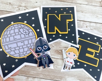 May include: A black and white banner with the word "ONE" in black letters with a yellow outline. The banner is decorated with silver stars on a black background. There are two small figures on the banner, one of Darth Vader and one of Luke Skywalker.