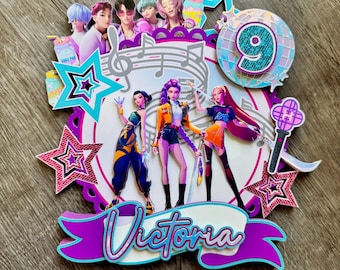 May include: A colorful cake topper featuring a pop music theme. The design includes illustrations of pop stars, musical notes, stars, and the number 9. The name "Victoria" is displayed on a banner.