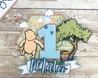 Pode incluir: Um topo de bolo com tema do Winnie the Pooh com um número um azul, um pote de mel, uma árvore e um personagem do Winnie the Pooh. O topo também inclui uma faixa com o nome "Mateo" escrito em azul.