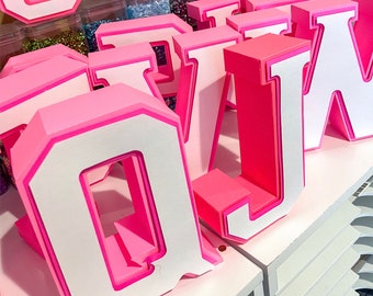 Puede incluir: Letras de cartón rosa y blanco que deletrean la palabra "JAW" con una letra "Q" rosa y blanca delante de la palabra. Las letras están sobre una superficie blanca.