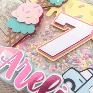 May include: A pink and white cake topper with the name "Arella" and the number "7" in the center. The cake topper is decorated with ice cream cones, sprinkles, and a blue truck.