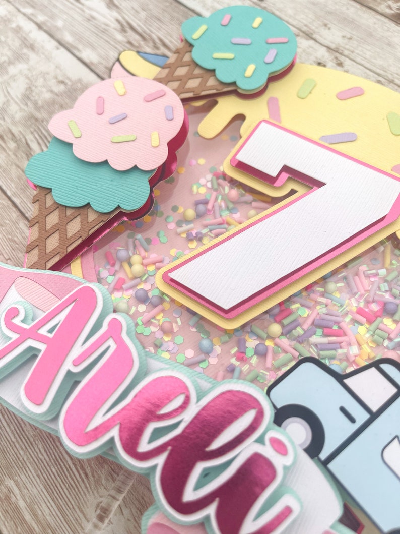 May include: A pink and white cake topper with the name "Arella" and the number "7" in the center. The cake topper is decorated with ice cream cones, sprinkles, and a blue truck.