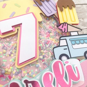 May include: A pink and white cake topper with a number 7, a blue ice cream truck, and the name "Arel" in pink letters. The cake topper is decorated with sprinkles and ice cream cones.