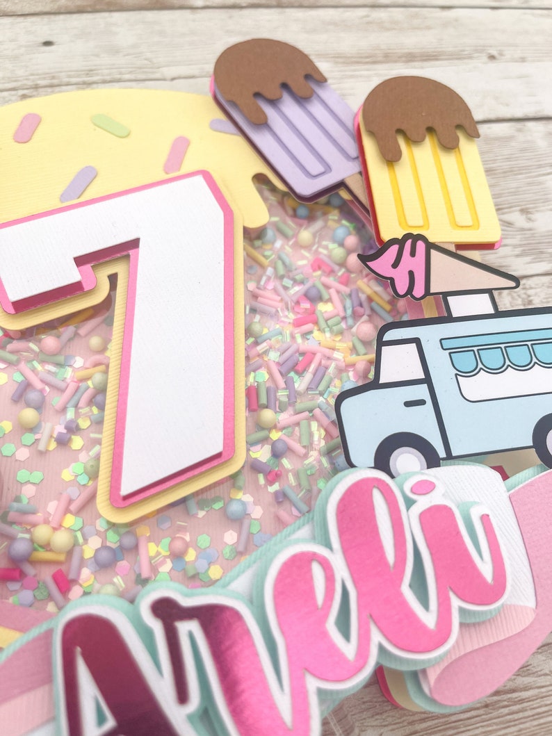 May include: A pink and white cake topper with a number 7, a blue ice cream truck, and the name "Arel" in pink letters. The cake topper is decorated with sprinkles and ice cream cones.
