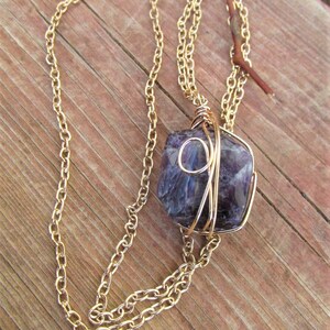 Puede incluir: Un collar de cadena dorada con una piedra de amatista p&uacute;rpura envuelta en alambre dorado. El collar est&aacute; sobre una superficie de madera.