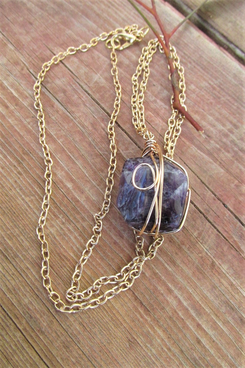 Puede incluir: Un collar de cadena dorada con una piedra de amatista p&uacute;rpura envuelta en alambre dorado. El collar est&aacute; sobre una superficie de madera.