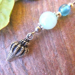 May include: A silver seashell charm hangs from a necklace with a light blue bead and a round, pale green bead. The necklace is on a textured, reddish-brown surface, with a green leaf in the background.