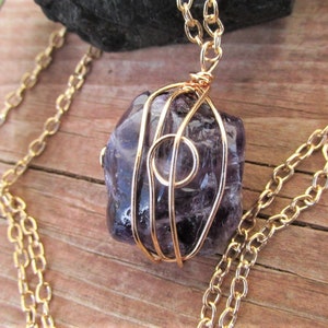 Puede incluir: Un collar de cadena dorada con una piedra de amatista p&uacute;rpura envuelta en alambre dorado. La piedra tiene una forma irregular y una textura natural y rugosa.