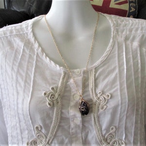 Puede incluir: Un collar de cadena dorada con un colgante de piedra p&uacute;rpura. El colgante est&aacute; envuelto en alambre dorado y tiene una forma org&aacute;nica &uacute;nica. El collar se lleva sobre una blusa blanca con ribetes decorativos.