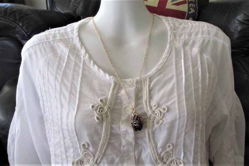 Puede incluir: Un collar de cadena dorada con un colgante de piedra p&uacute;rpura. El colgante est&aacute; envuelto en alambre dorado y tiene una forma org&aacute;nica &uacute;nica. El collar se lleva sobre una blusa blanca con ribetes decorativos.