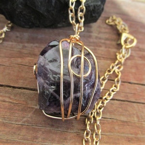Puede incluir: Un collar de cadena dorada con una piedra de amatista p&uacute;rpura envuelta en alambre dorado. La piedra tiene una forma irregular y una textura natural y rugosa.