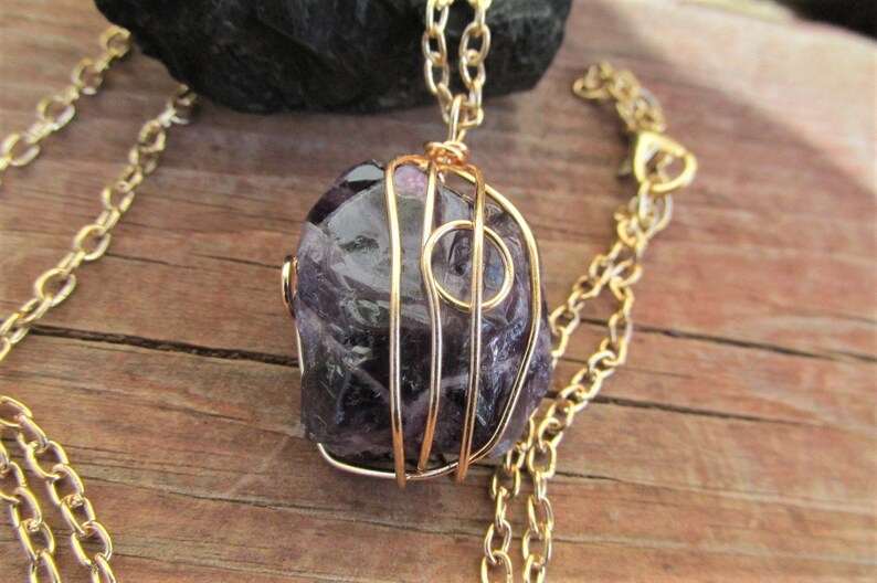 Puede incluir: Un collar de cadena dorada con una piedra de amatista p&uacute;rpura envuelta en alambre dorado. La piedra tiene una forma irregular y una textura natural y rugosa.